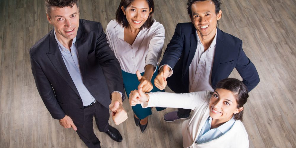 Four smiling middle-aged business people standing in office hall, looking at camera and showing thumbs up. High angle view.